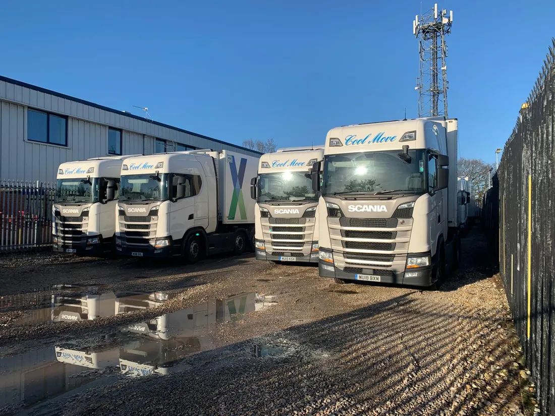A row of four white Cool Move Scania tractor units lined up nose-on against a clear blue sky, reflected in a yard puddle.